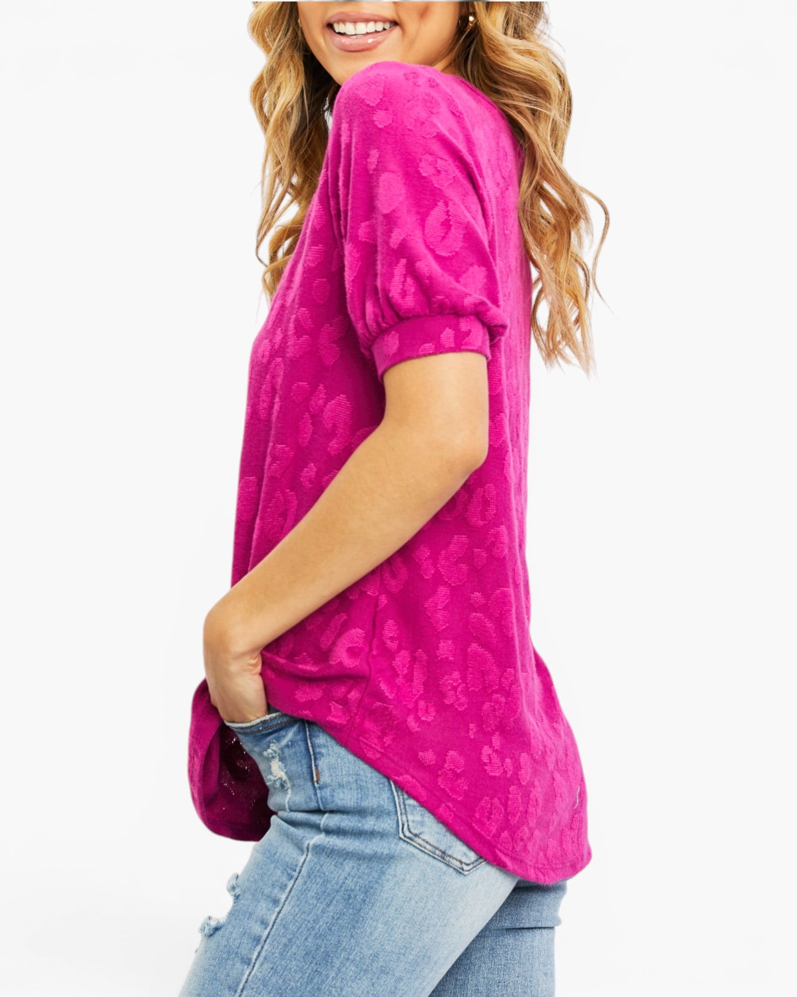 A woman wearing a bright pink short-sleeve top with a tonal paisley pattern, paired with light-wash blue jeans, shown from a side-back angle against a plain white background.