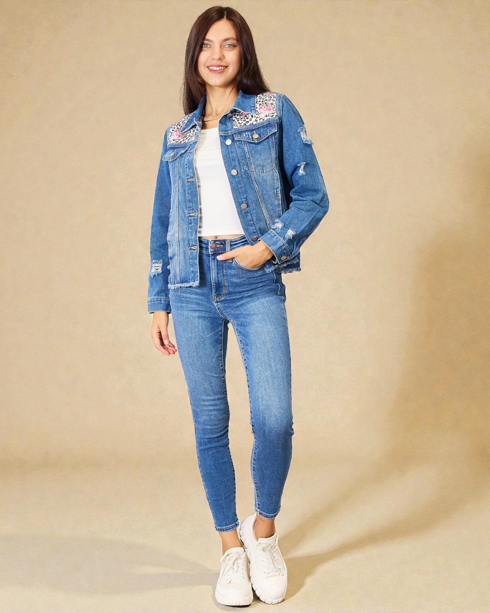 Woman wearing a blue denim jacket with floral embroidery and ripped details, paired with blue jeans and white sneakers on a beige background.