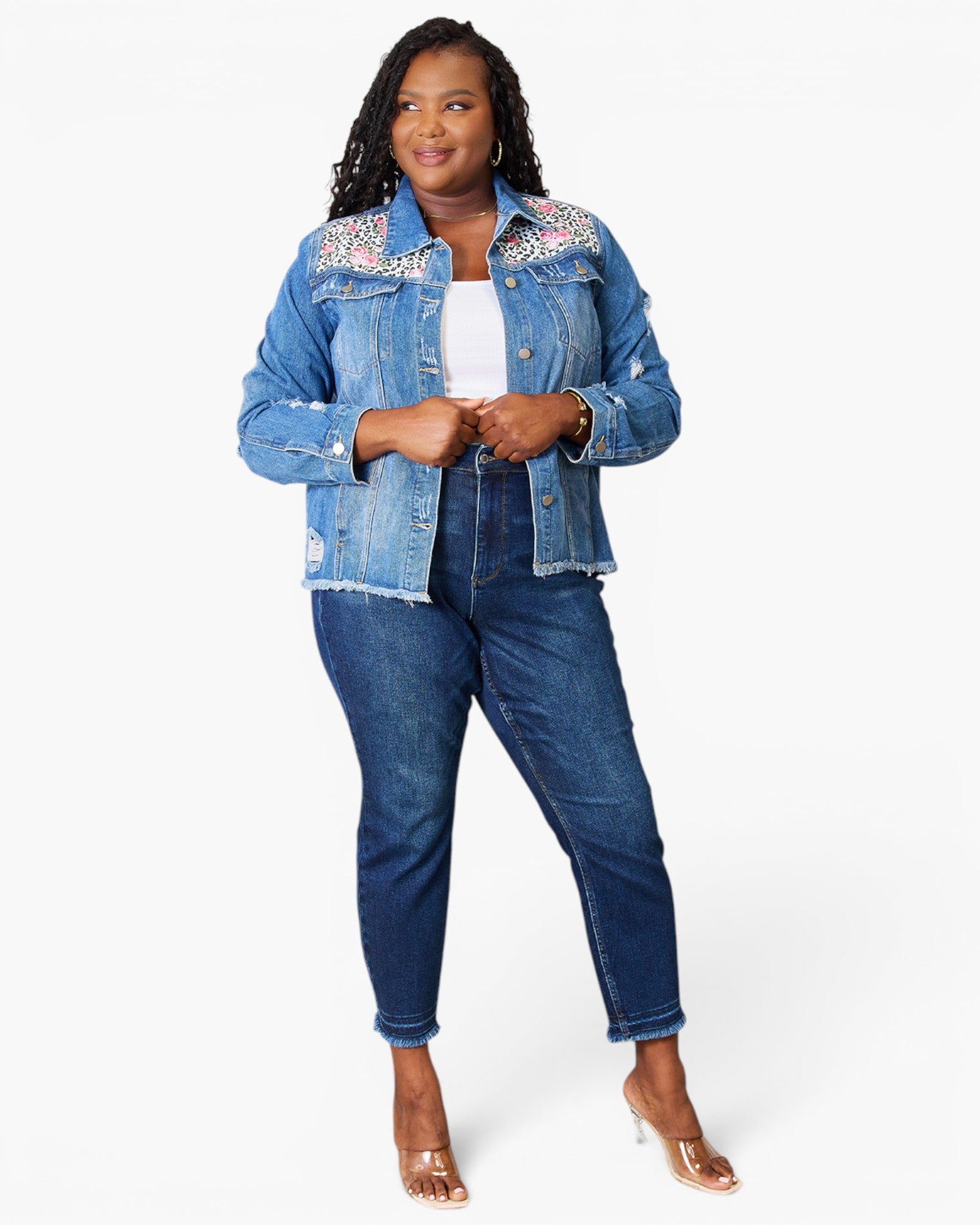 A woman modeling a light blue denim jacket with colorful floral embroidery on the shoulders, paired with dark blue jeans and clear high-heeled sandals, against a plain white background.