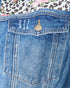 Close-up of the back pocket of blue denim jeans, showing a single gold-tone button, distressed stitching, and contrast stitching. A floral and leopard print fabric is visible at the top edge.