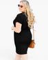 A woman modeling a fitted black short-sleeve bodycon dress, shown from a side-back angle against a plain white studio background. She is wearing round sunglasses and carrying a small brown crossbody bag.