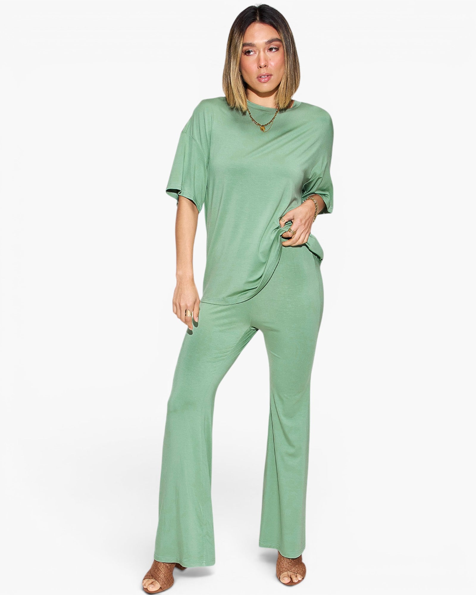 A woman modeling a sage green short-sleeve jumpsuit with a draped neckline and wide-leg pants, standing against a plain white studio background.