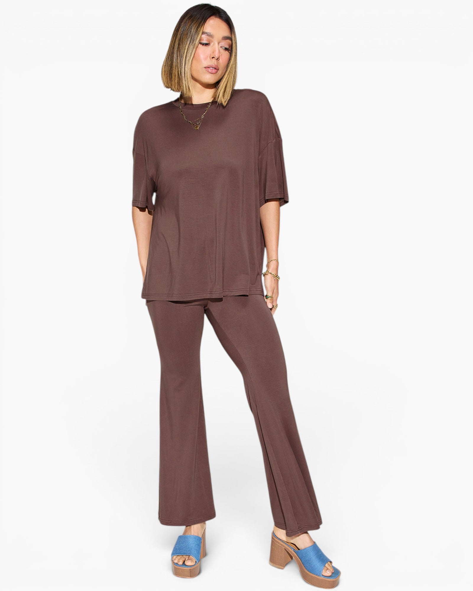 A woman modeling a matching two-piece lounge set in a solid dark brown color, featuring a short-sleeve top and wide-leg pants, paired with blue platform sandals against a plain white background.