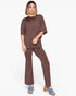 A woman modeling a matching brown lounge set consisting of a short-sleeve top and wide-leg pants, paired with blue platform sandals, against a plain white studio background.