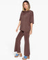 A woman modeling a matching brown lounge set consisting of a short-sleeve top and wide-leg pants, paired with blue platform slide sandals, against a plain white background.