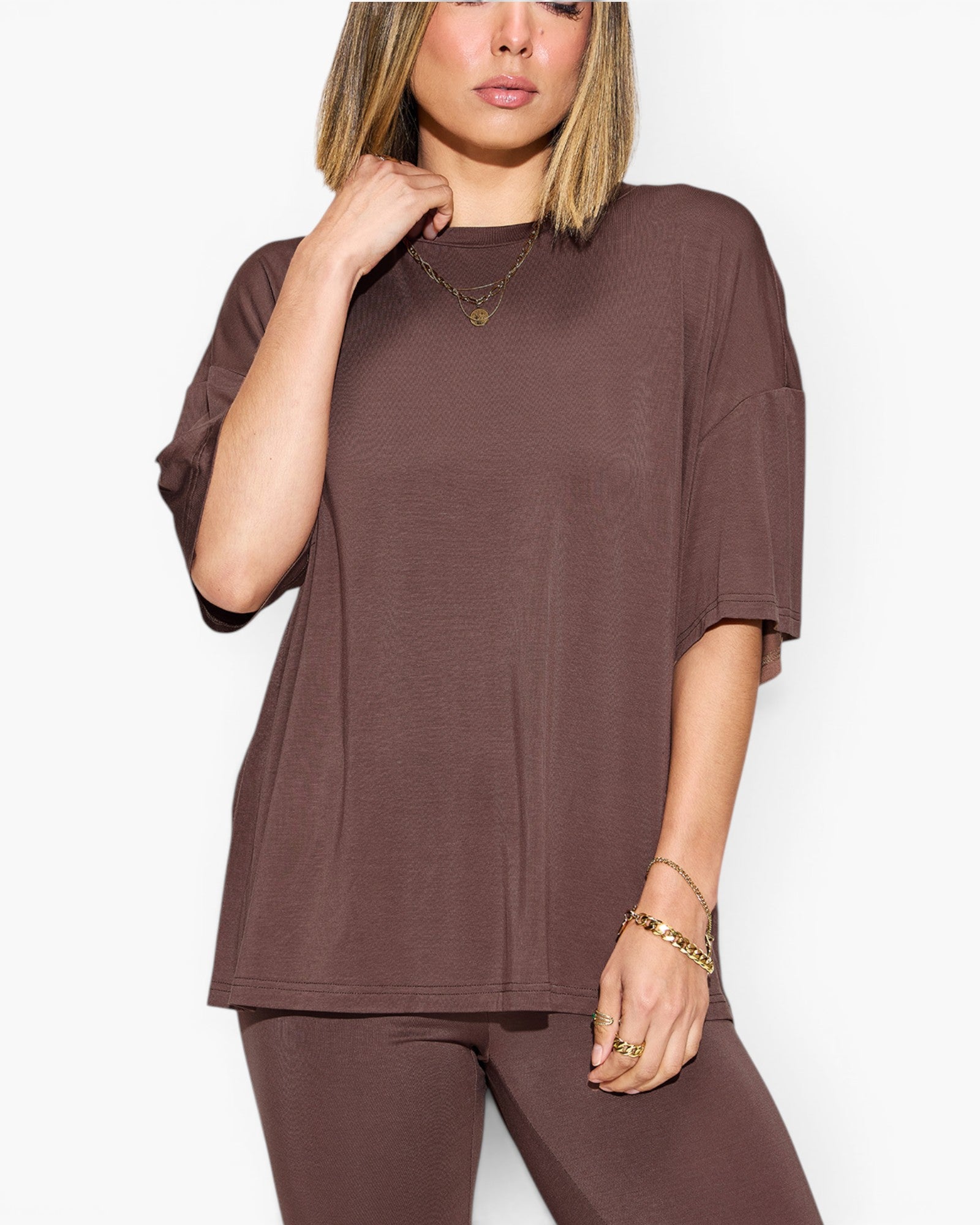 A woman wearing an oversized short-sleeve t-shirt and matching jogger pants in a solid dark brown color, styled with gold jewelry against a plain white background.