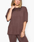 A woman wearing an oversized short-sleeve t-shirt and matching jogger pants in a solid dark brown color, styled with gold jewelry against a plain white background.