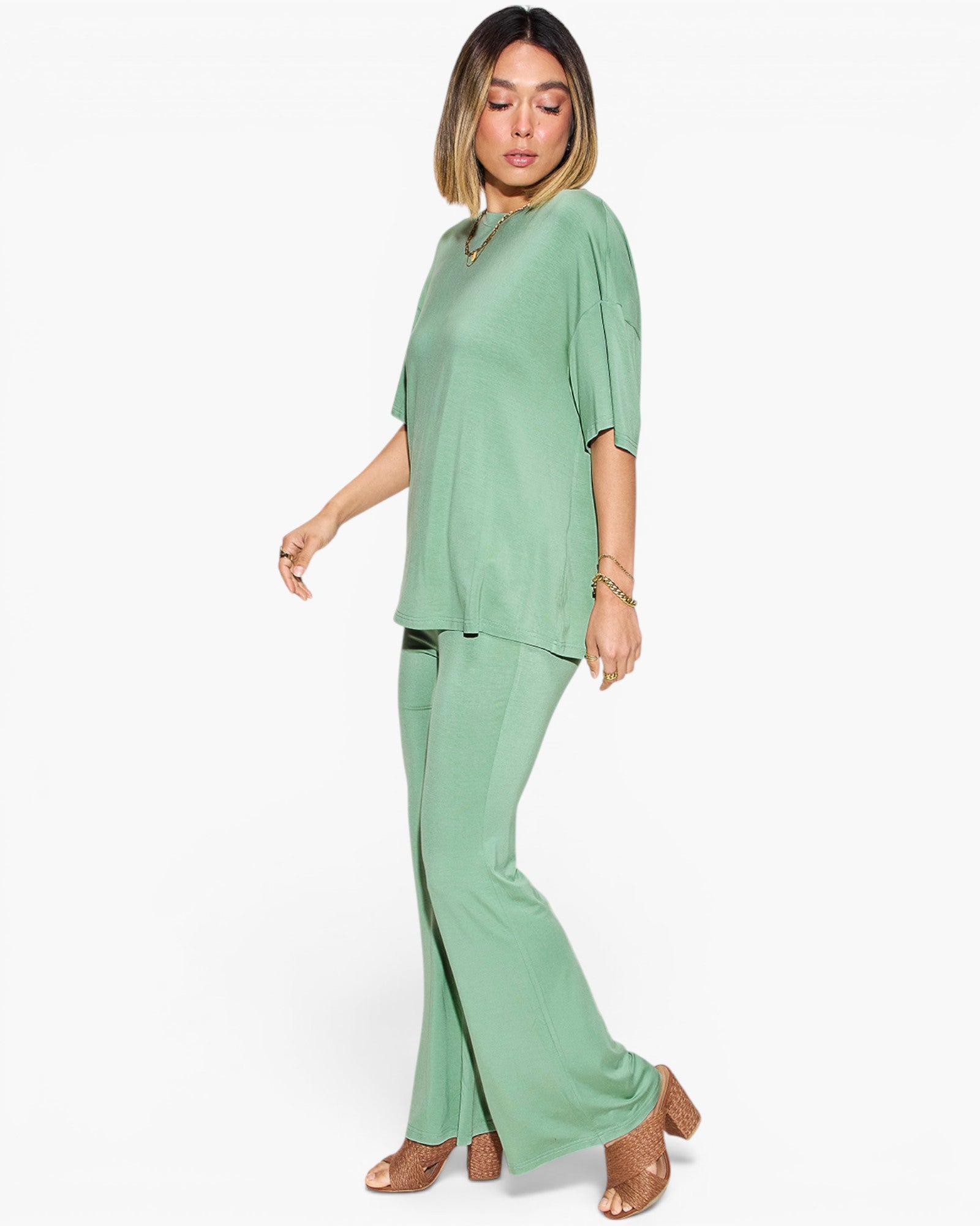 A woman modeling a sage green two-piece lounge set consisting of a short-sleeve V-neck top and matching wide-leg pants, paired with tan heeled sandals against a plain white background.