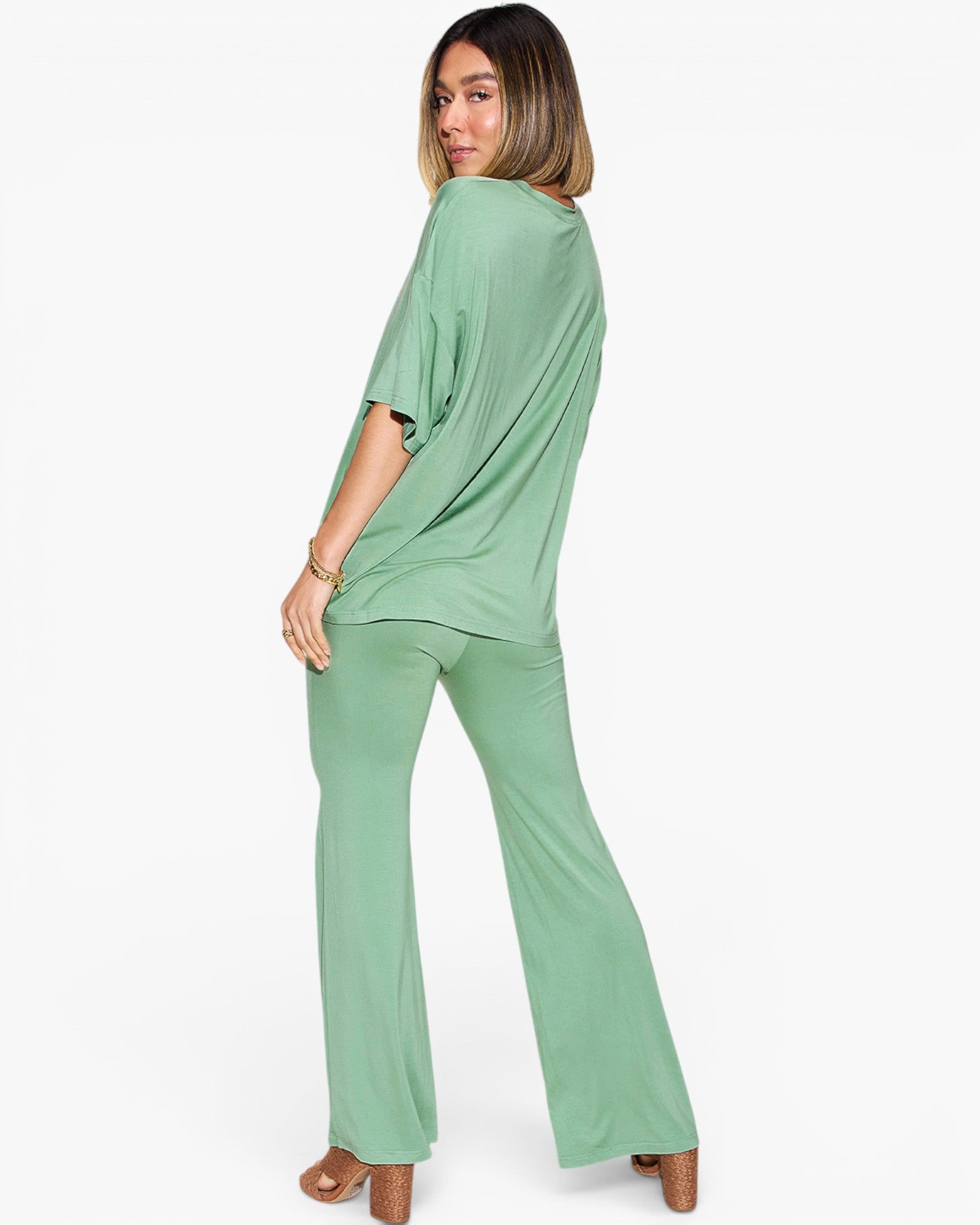 A woman modeling a sage green two-piece lounge set consisting of a short-sleeve draped top and matching wide-leg pants, shown from a three-quarters back view against a plain white background.