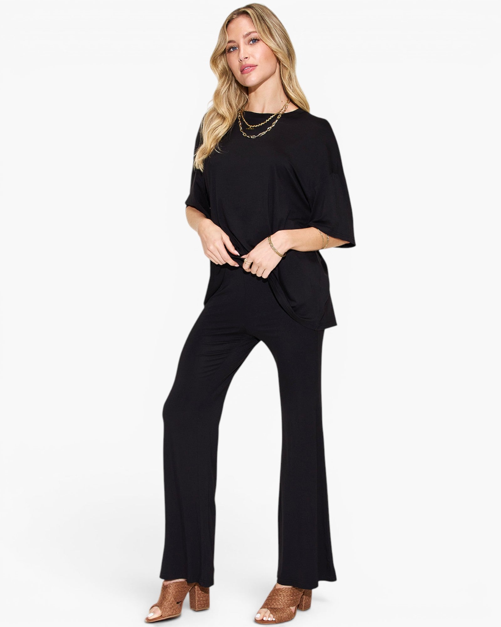 A woman modeling a black short-sleeve top and matching black wide-leg pants, styled with layered gold necklaces and brown heeled sandals against a plain white studio background.