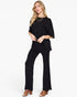 A woman modeling a black short-sleeve top and matching black wide-leg pants, styled with layered gold necklaces and brown heeled sandals against a plain white studio background.