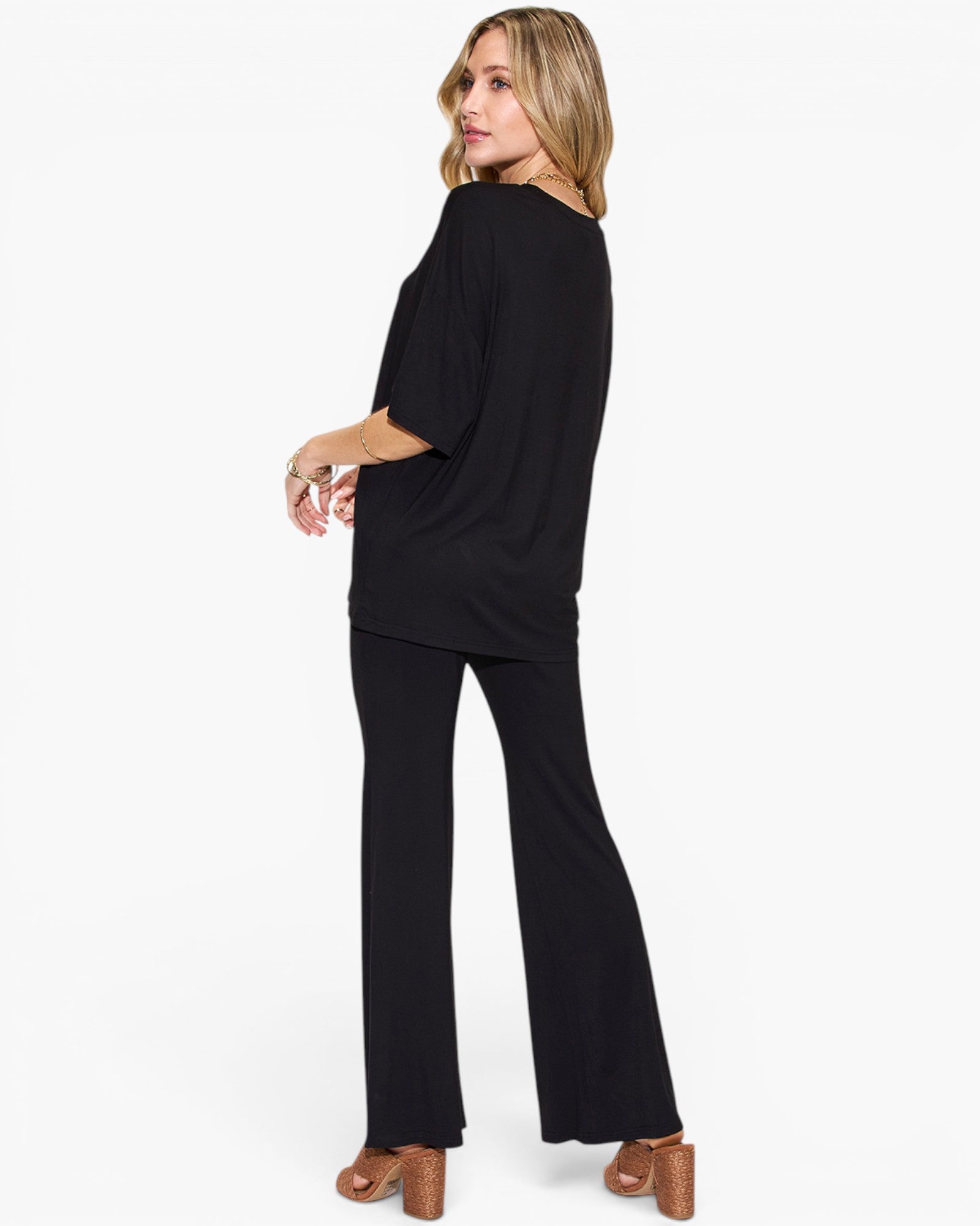 A woman modeling a black two-piece outfit consisting of a short-sleeve draped top and matching wide-leg pants, shown from a side-back angle against a plain white studio background.