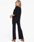 A woman modeling a black two-piece outfit consisting of a short-sleeve draped top and matching wide-leg pants, shown from a side-back angle against a plain white studio background.