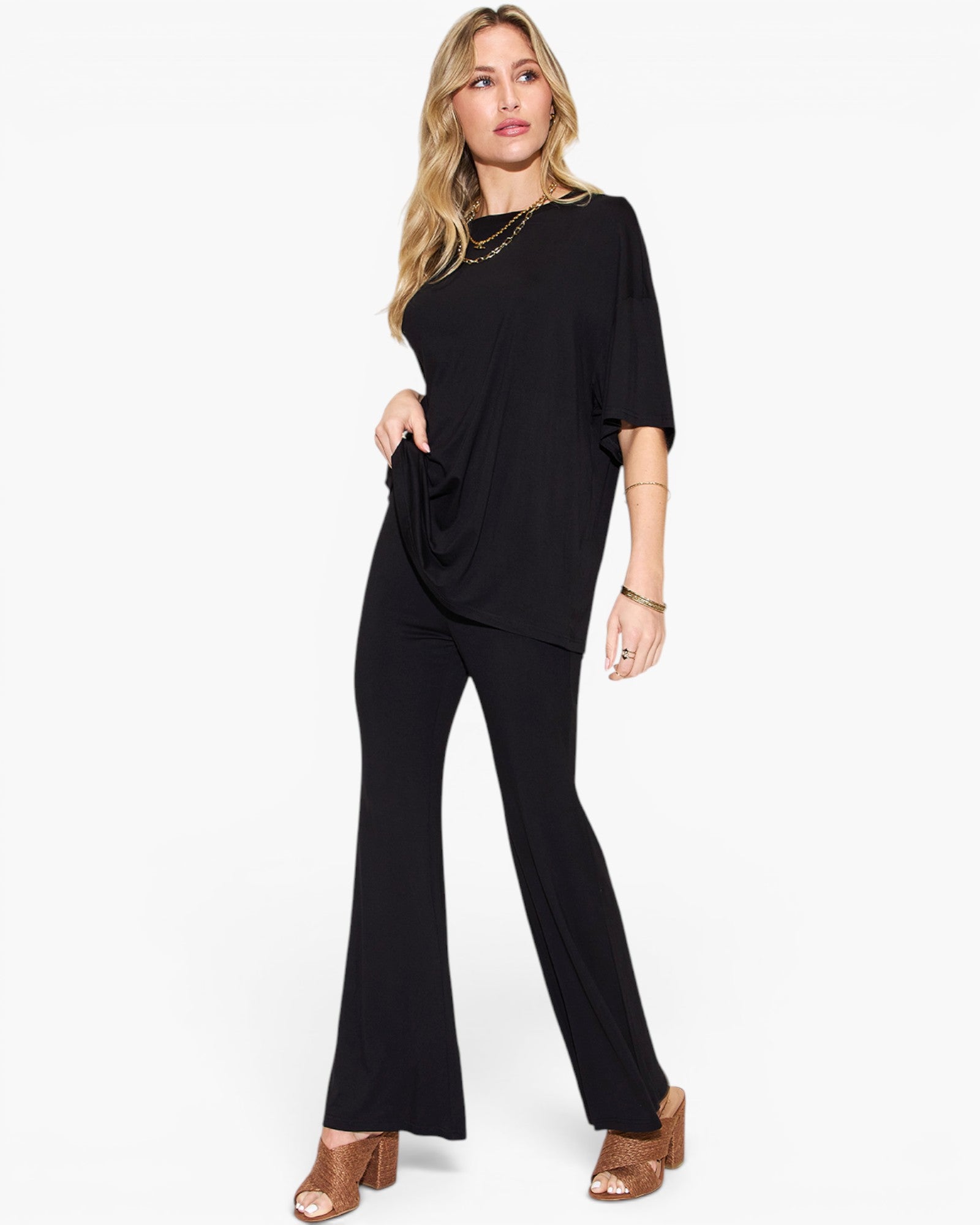 A woman modeling a black draped short-sleeve jumpsuit with flared legs, styled with gold jewelry and brown heeled sandals against a plain white background.