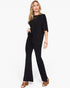 A woman modeling a black draped short-sleeve jumpsuit with flared legs, styled with gold jewelry and brown heeled sandals against a plain white background.