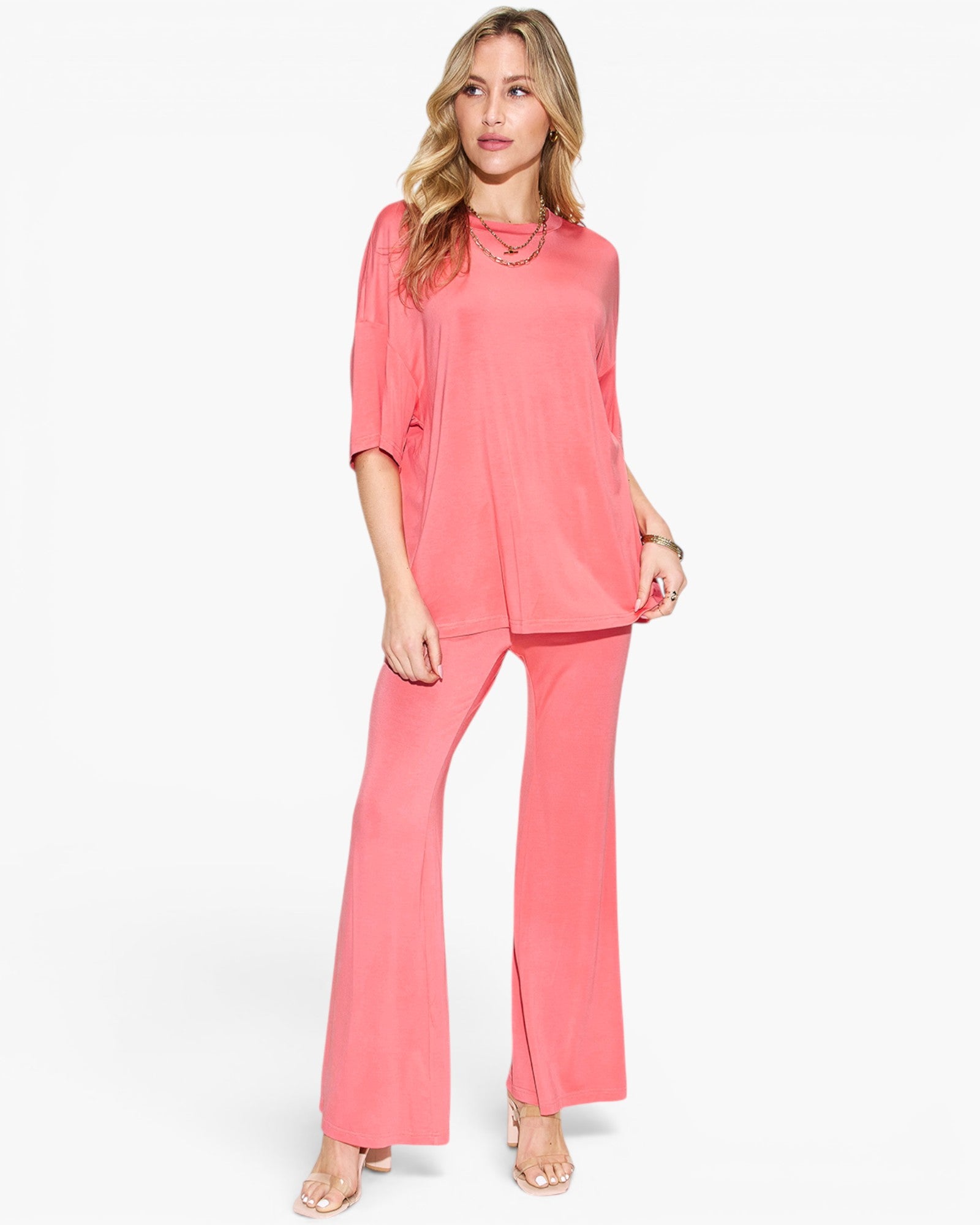 A woman modeling a coral pink two-piece set consisting of a short-sleeve top and matching wide-leg pants, photographed against a clean white studio background.