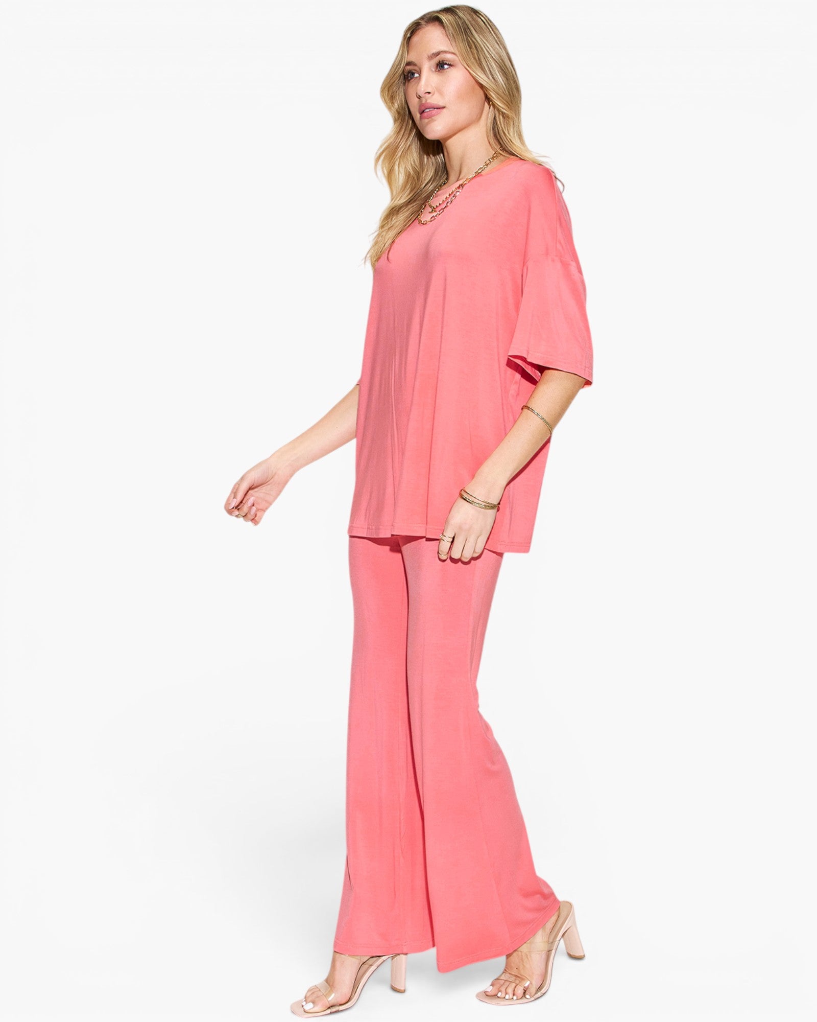 A woman modeling a two-piece coral pink lounge set consisting of a short-sleeve top and matching wide-leg pants, paired with clear high-heeled sandals against a plain white background.