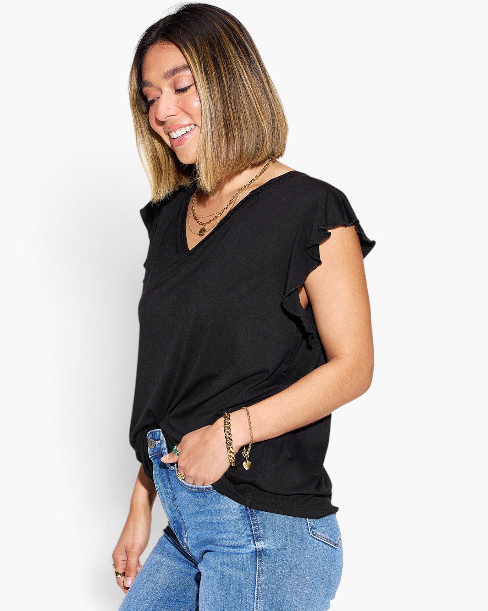 A woman wearing a black short-sleeve V-neck top with ruffled sleeve edges, paired with blue jeans, photographed against a plain light gray studio background.