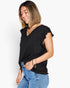 A woman wearing a black short-sleeve V-neck top with ruffled sleeve edges, paired with blue jeans, photographed against a plain light gray studio background.
