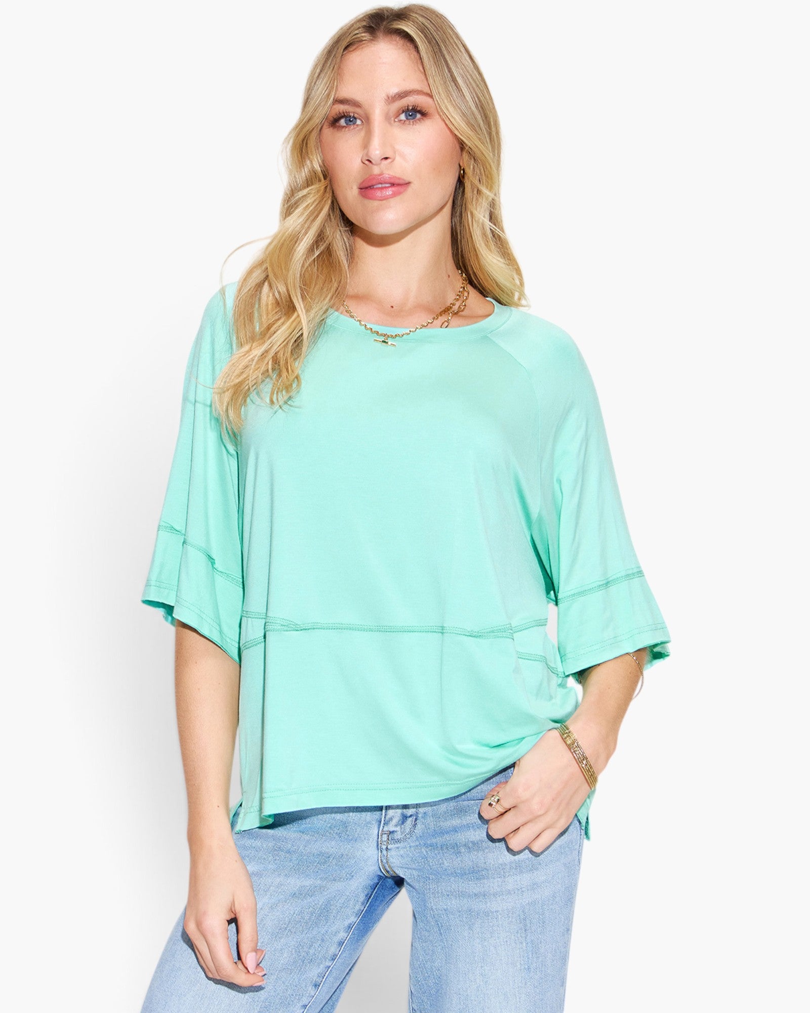 A woman wearing a mint green short-sleeve top with layered ruffle details on the sleeves and a horizontal seam near the waist, paired with light blue jeans, against a plain white background.