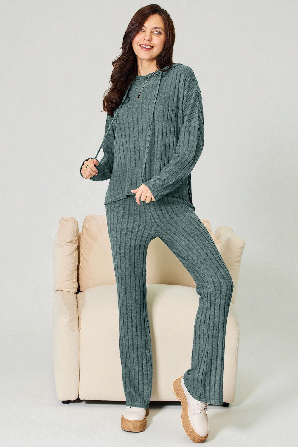 Woman wearing a green ribbed sweater and pants sitting on a beige chair against a white background