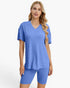 A woman modeling a matching blue ribbed lounge set consisting of a short-sleeve V-neck top and fitted biker shorts against a plain light gray studio background.