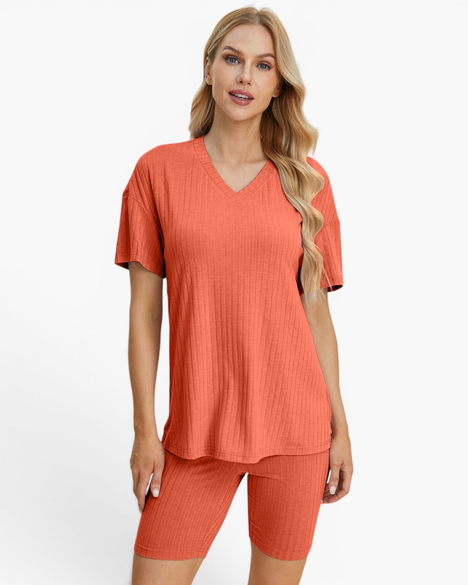 A woman modeling a matching two-piece lounge set consisting of a short-sleeve V-neck top and ribbed biker shorts in a warm coral-orange color, set against a plain light gray background.