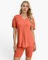 A woman modeling a matching two-piece lounge set consisting of a short-sleeve V-neck top and ribbed biker shorts in a warm coral-orange color, set against a plain light gray background.