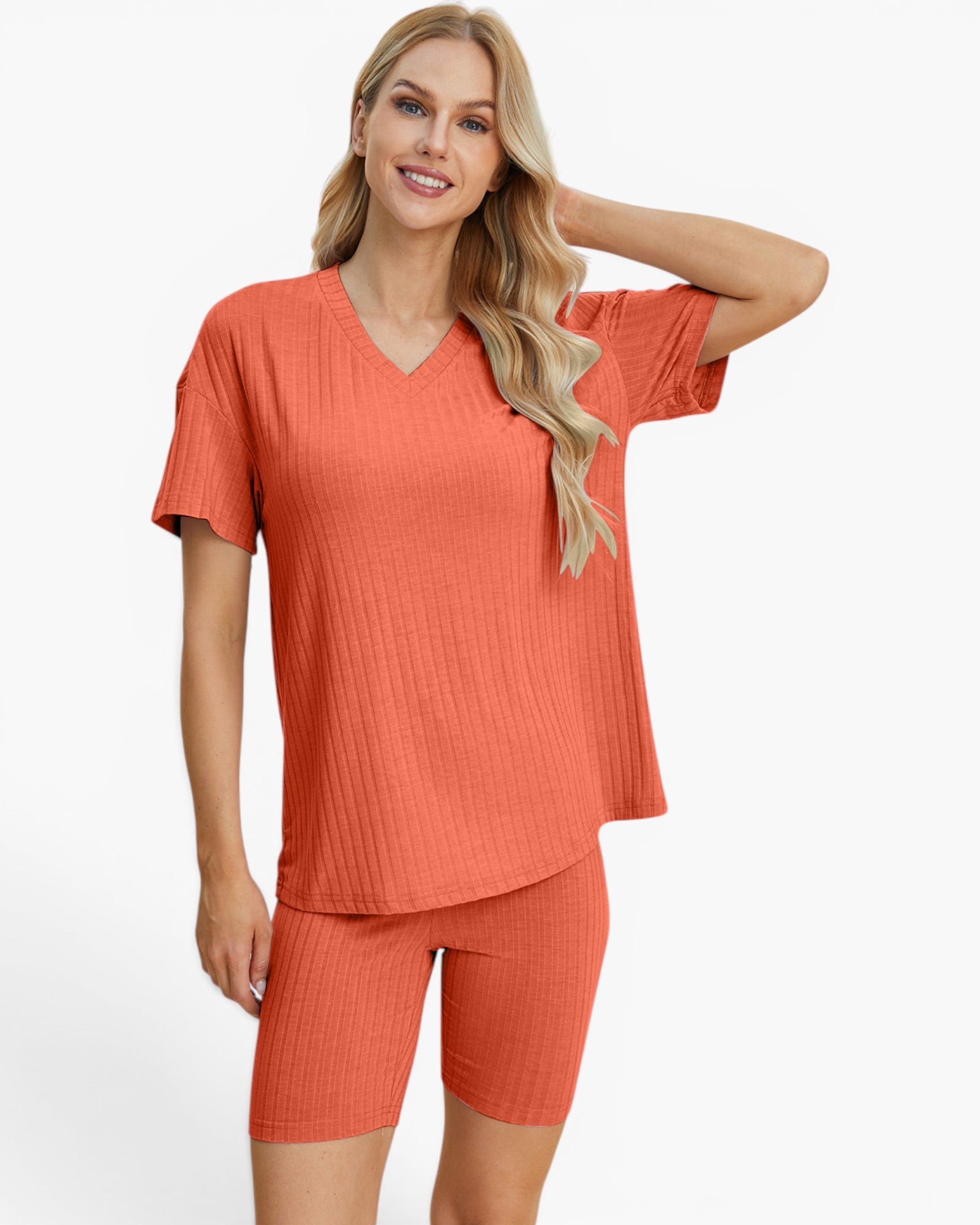 A woman modeling a matching two-piece ribbed knit loungewear set in a warm coral-orange color, featuring a short-sleeve V-neck top and matching biker shorts, against a plain white studio background.