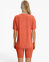 A woman shown from the back wearing a matching ribbed short-sleeve top and biker shorts set in a warm coral-orange color, against a plain white studio background.
