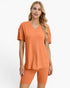 A woman modeling a matching two-piece lounge set consisting of a short-sleeve V-neck ribbed top and matching ribbed biker shorts in a warm orange color, set against a plain white studio background.