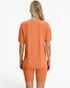 A woman shown from the back wearing a matching ribbed short-sleeve top and biker shorts set in a warm orange color, against a plain white studio background.