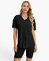 A woman modeling a black short-sleeve V-neck top with a subtle vertical ribbed texture, paired with matching black bike shorts, against a plain white studio background.
