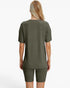 A woman viewed from the back wearing a ribbed short-sleeve top and matching ribbed biker shorts in an olive green color, set against a plain white studio background.