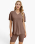 A woman modeling a matching ribbed lounge set consisting of a short-sleeve V-neck top and matching biker shorts in a warm taupe-brown color, set against a plain white studio background.