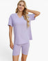 A woman modeling a light purple ribbed two-piece pajama set consisting of a short-sleeve V-neck top and matching fitted shorts, against a plain white studio background.