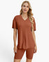 A woman modeling a matching rust-colored ribbed lounge set consisting of a short-sleeve V-neck top and fitted biker shorts, against a plain white studio background.
