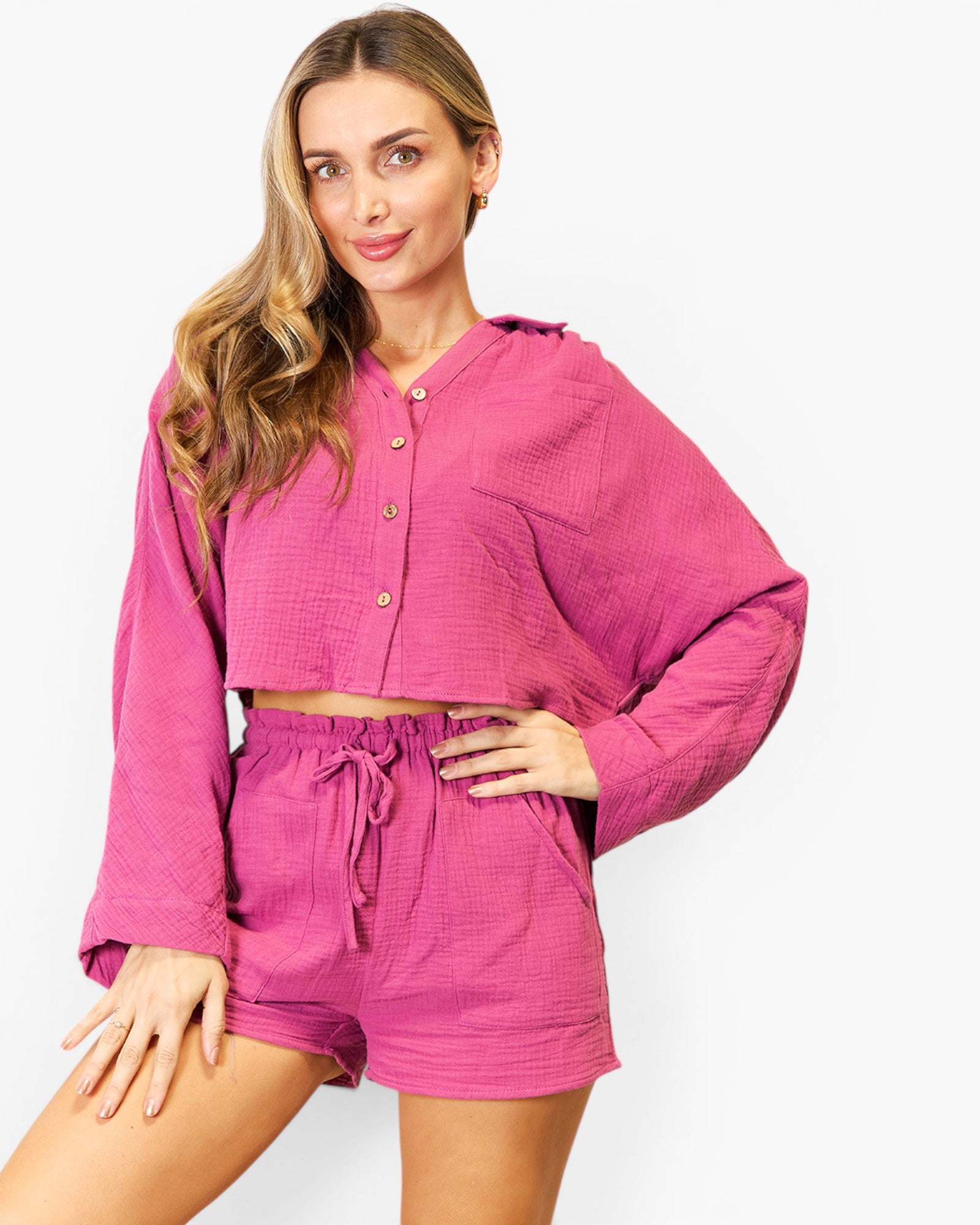 A woman modeling a matching two-piece lounge set in a textured magenta pink fabric, featuring a cropped long-sleeve button-up shirt and coordinating drawstring shorts, set against a plain light gray background.