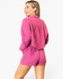 A woman wearing a bright pink textured two-piece set consisting of a long-sleeve collared top and matching shorts, shown from a three-quarters back view against a plain white background.