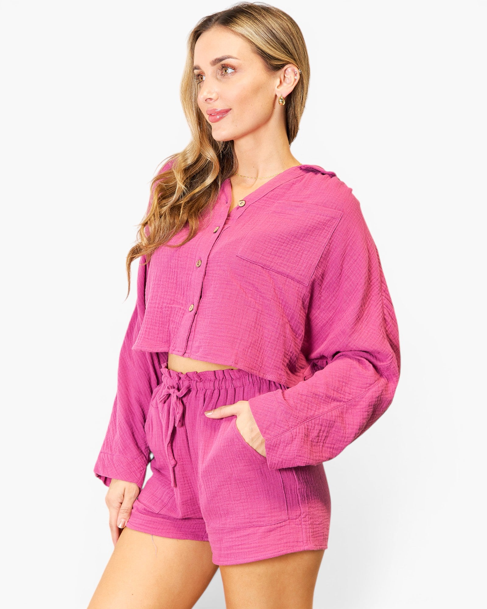 A woman modeling a matching two-piece lounge set in a textured magenta pink fabric, featuring a cropped button-front long-sleeve top and coordinating shorts, against a plain white studio background.