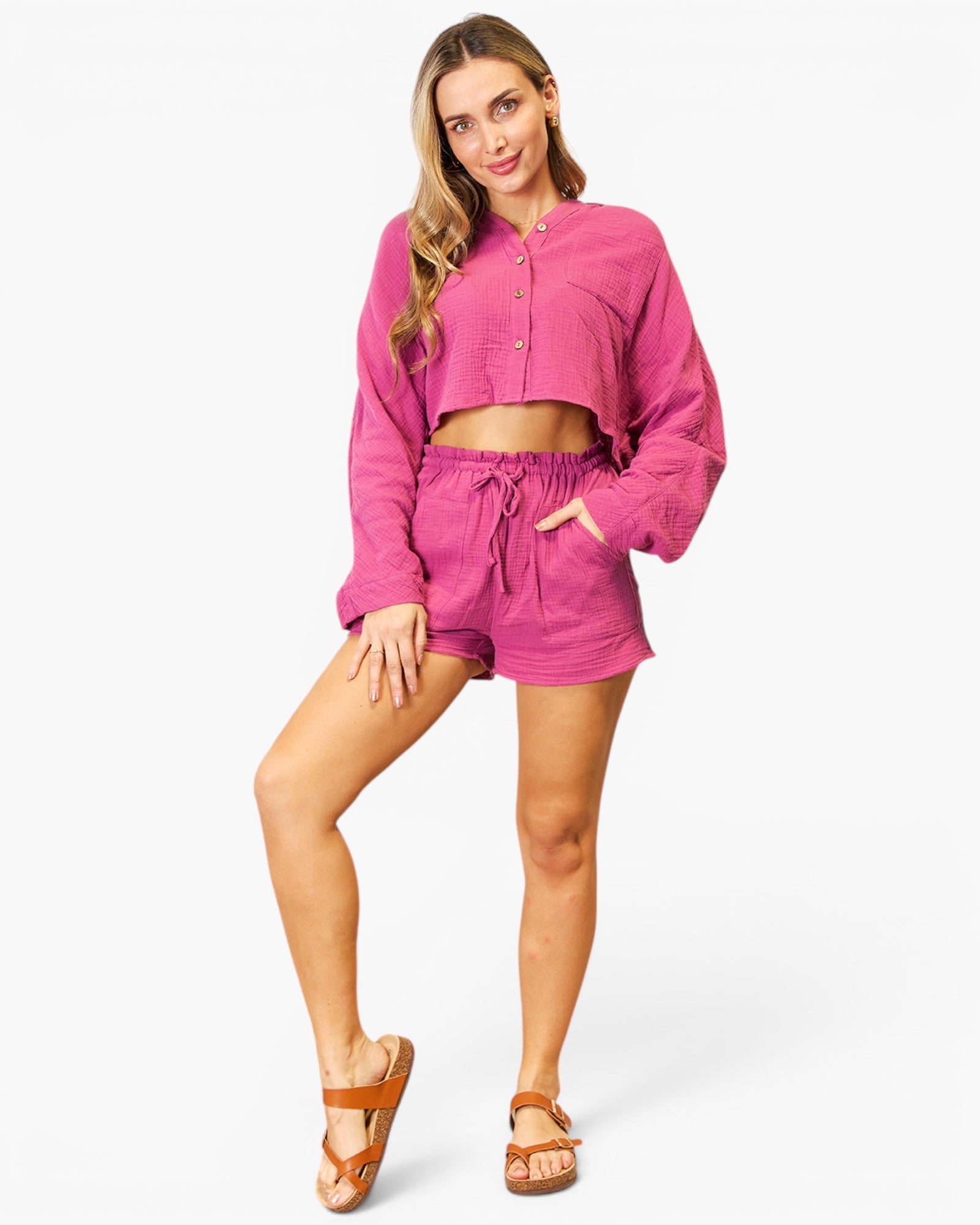 A woman modeling a matching two-piece lounge set in a vibrant magenta pink, featuring a cropped long-sleeve button-up shirt and coordinating drawstring shorts, paired with brown sandals against a light gray studio background.