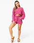 A woman modeling a matching two-piece lounge set in a vibrant magenta pink, featuring a cropped long-sleeve button-up shirt and coordinating drawstring shorts, paired with brown sandals against a light gray studio background.