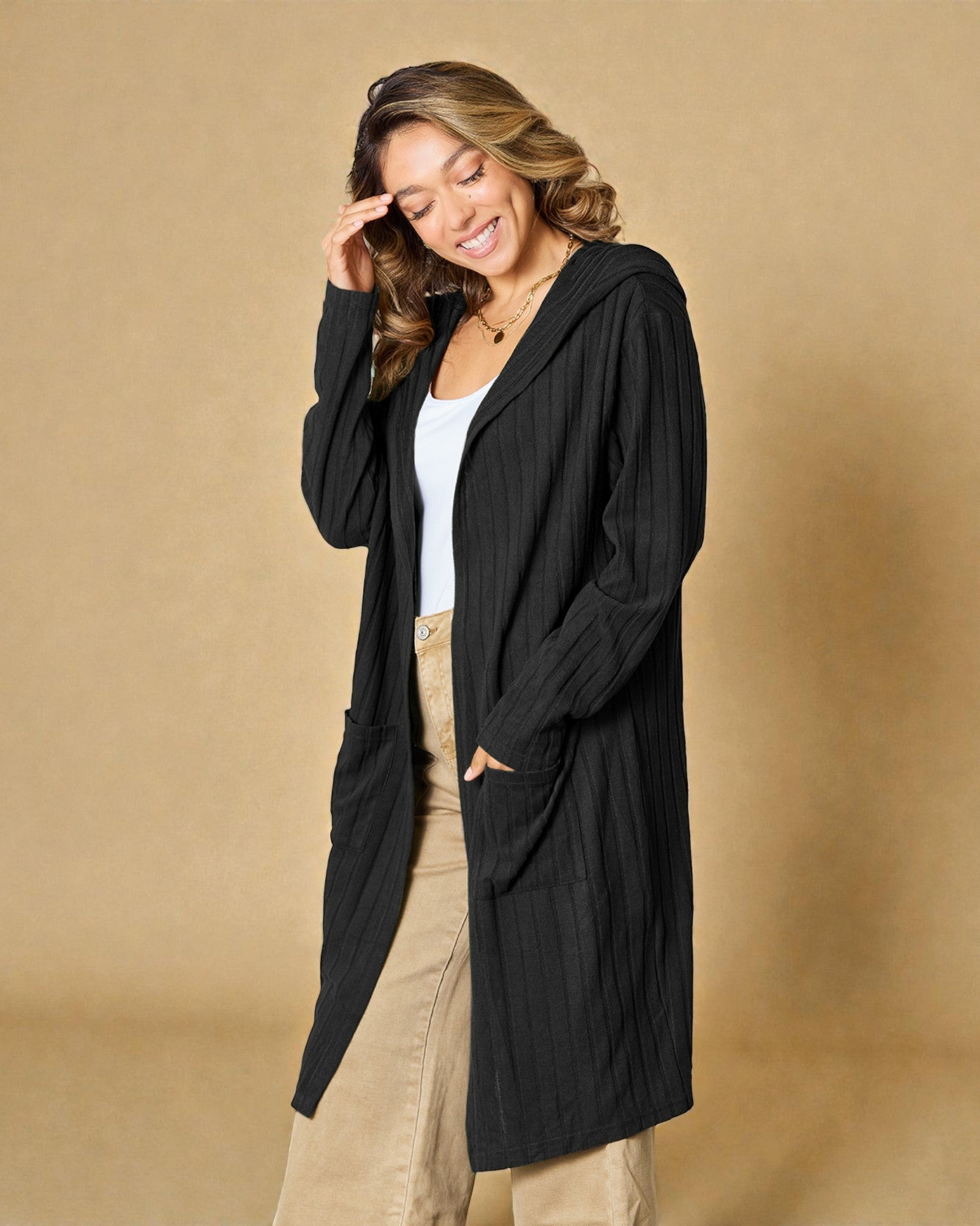 Woman wearing a black cardigan over a white top and beige pants on a beige background
