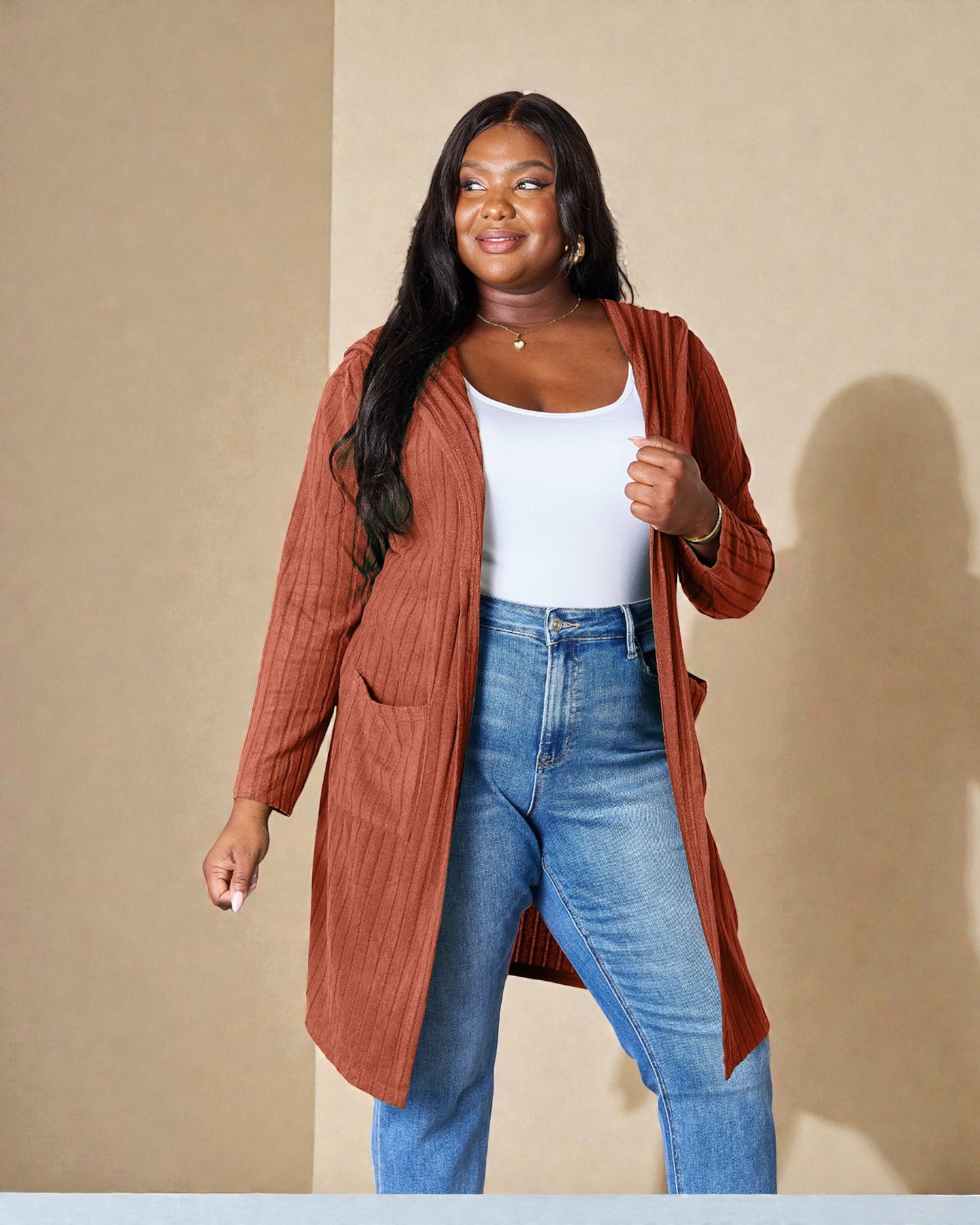 Woman wearing a rust-colored cardigan, white top, and blue jeans against a beige background