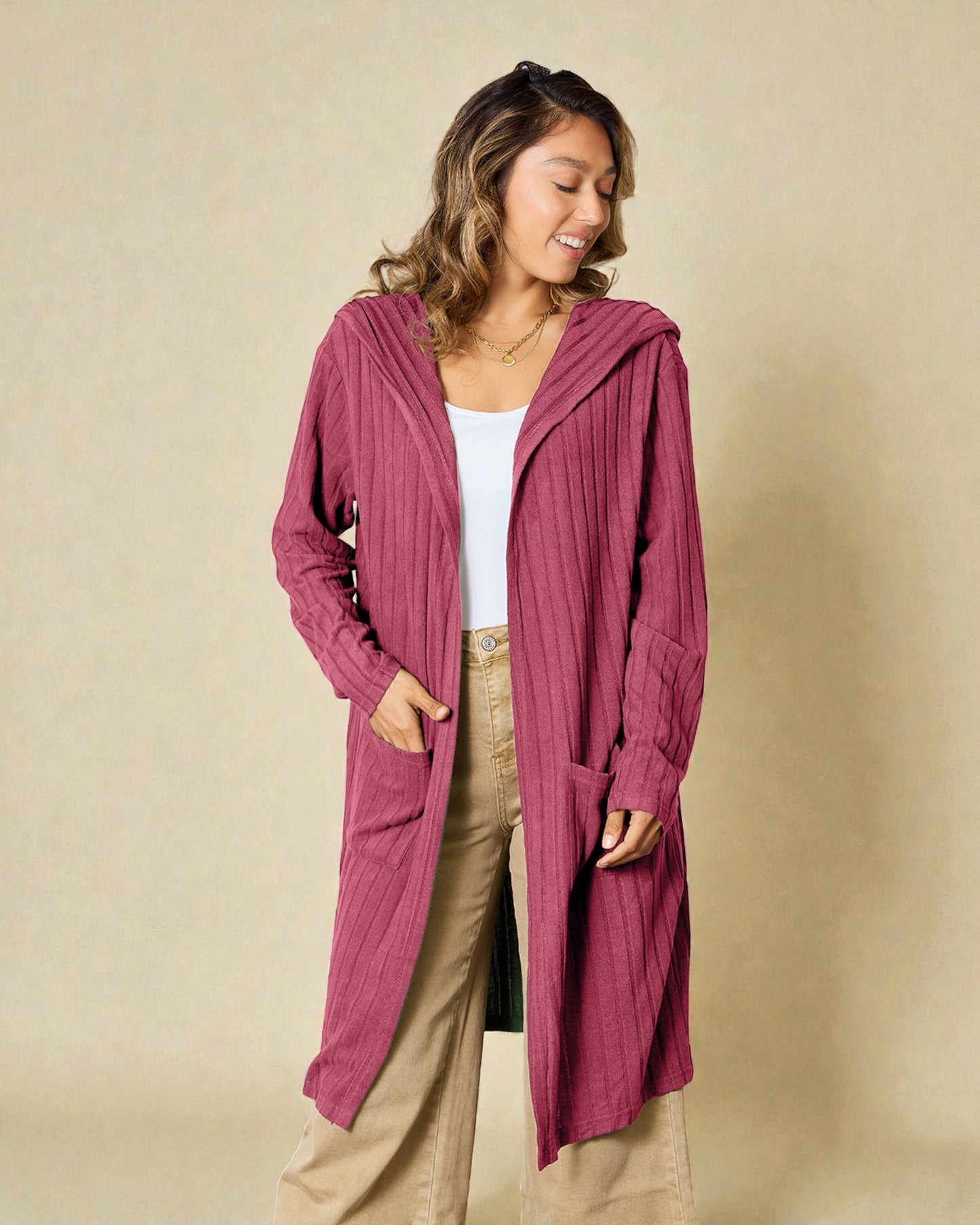 Woman wearing a long pink cardigan over a white top and beige pants on a beige background