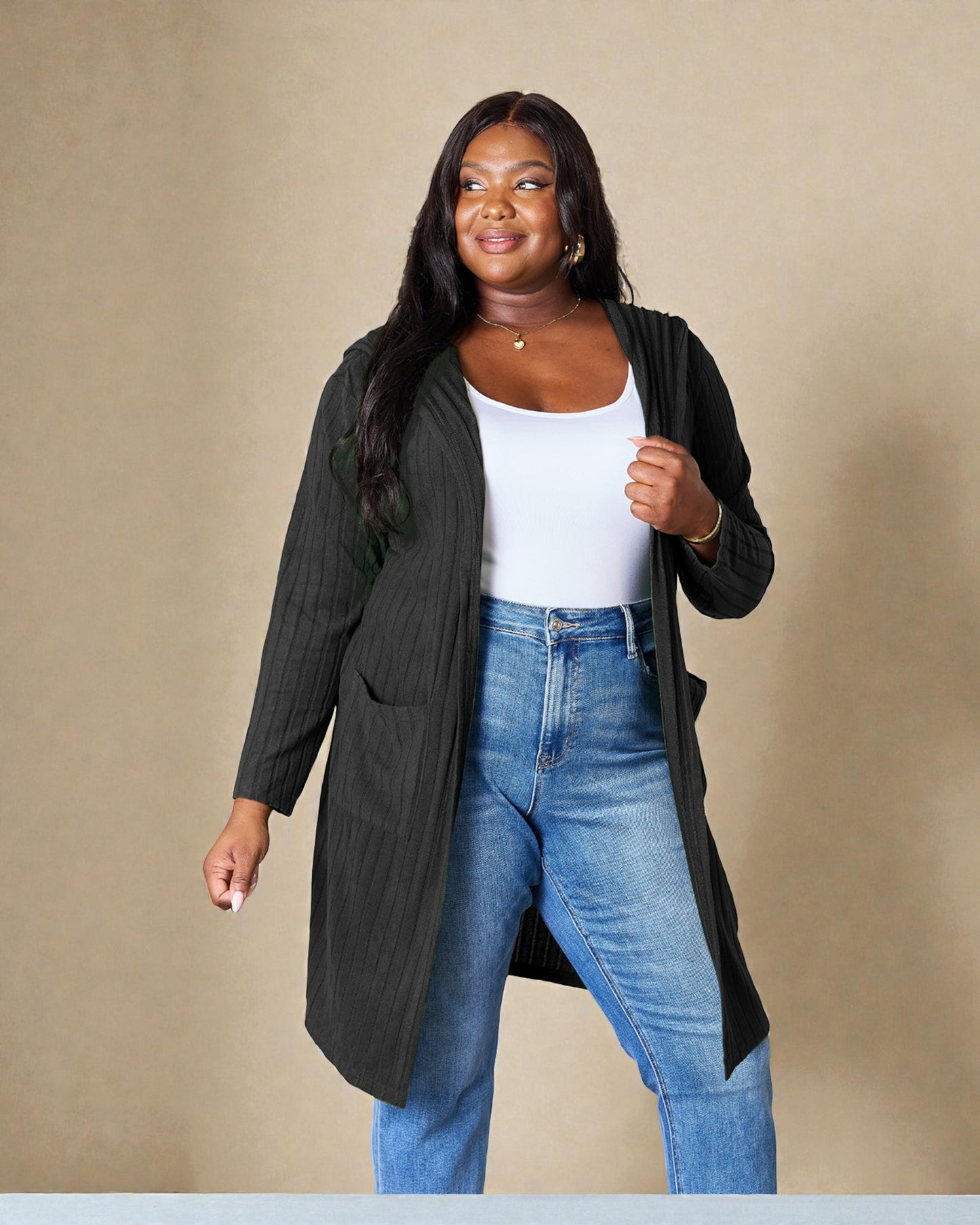 Woman wearing a long dark green cardigan over a white top and blue jeans on a beige background