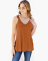 A woman modeling a rust-orange sleeveless tank top with a scalloped lace V-neckline, paired with blue jeans against a plain white background.