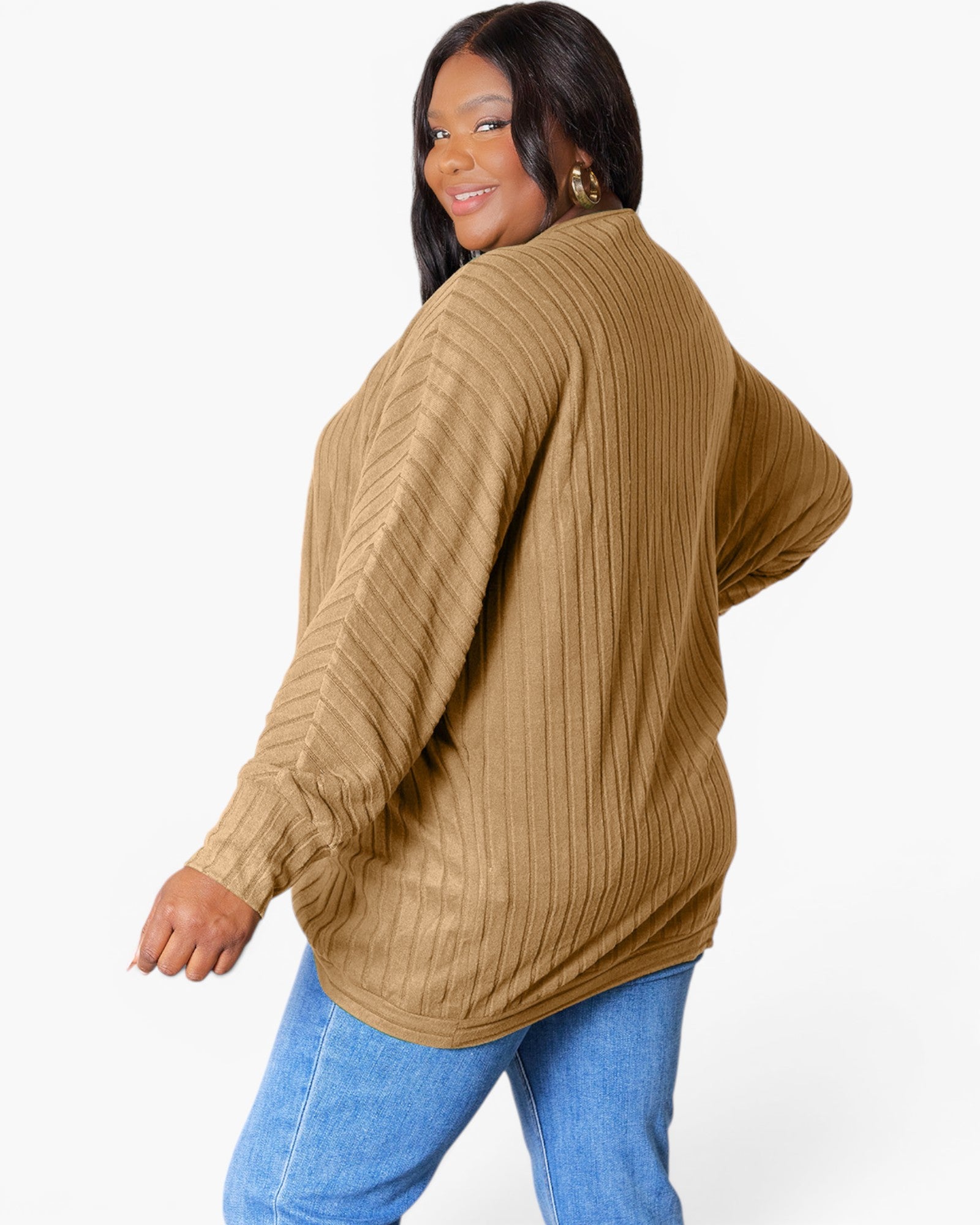 A woman wearing a camel brown ribbed knit sweater with blue jeans, photographed from a three-quarters back angle against a plain white studio background.