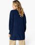 A woman shown from the back wearing a navy blue ribbed knit sweater and beige pants against a light gray studio background.