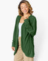 A woman wearing a dark green, long-sleeve, open-front cardigan with a textured crinkle fabric over a white tank top and beige pants, photographed against a clean white studio background.